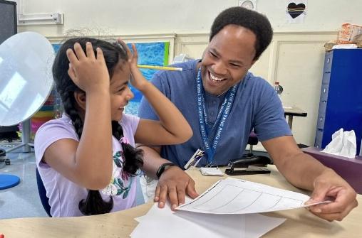  Christopher 车道 smiles as he holds a piece of paper while sitting next to a student.